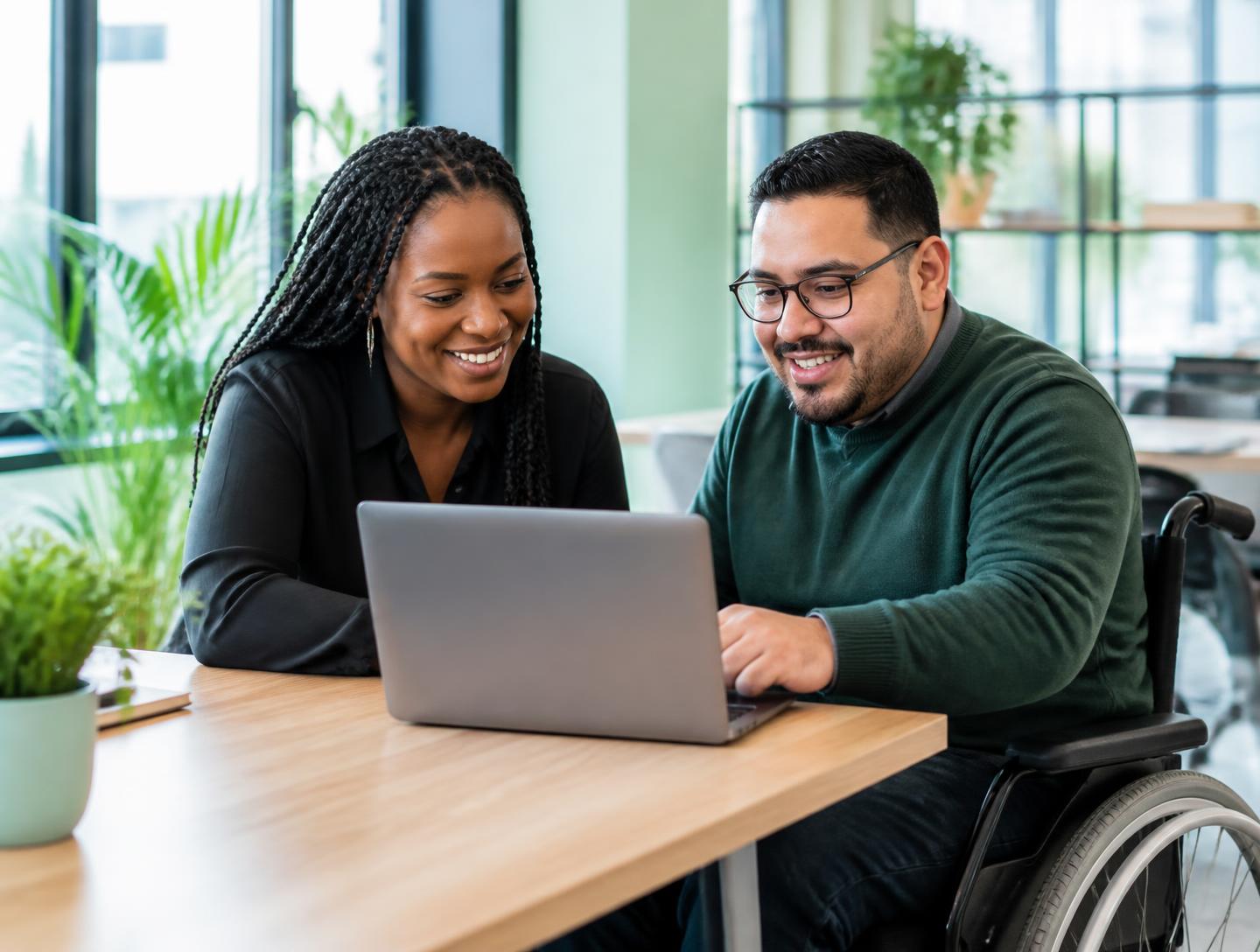 Two colleagues, one seated in a wheelchair, reviewing work together on a laptop in a bright office.